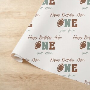 Op de afbeelding: Wit verpakkingspapier met een herhalend patroon van bruine en groene voetballen, het woord "ONE" in bruine en groene letters, en de tekst "Happy Birthday Aiden" en "year down".