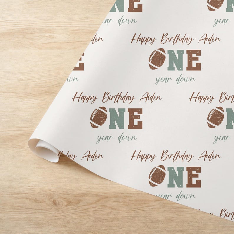 Op de afbeelding: Wit verpakkingspapier met een herhalend patroon van bruine en groene voetballen, het woord "ONE" in bruine en groene letters, en de tekst "Happy Birthday Aiden" en "year down".