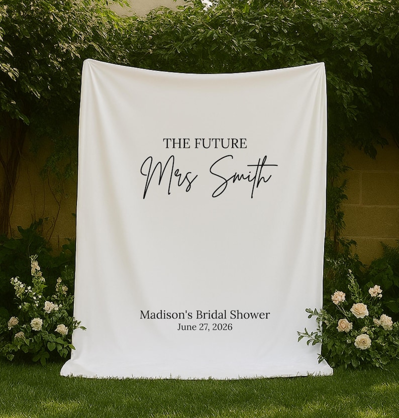 May include: A white fabric backdrop with the text "THE FUTURE Mrs Smith" in elegant black script. Below, it reads "Madison's Bridal Shower June 27, 2026". The backdrop is set outdoors with greenery and flowers.