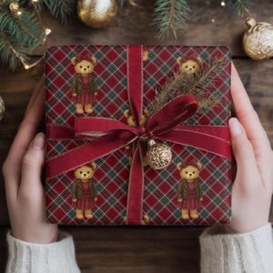 May include: A wrapped gift with red plaid wrapping paper featuring teddy bears in dresses. The gift is tied with a burgundy ribbon and a gold ornament. The gift is held by a person wearing a white sweater.