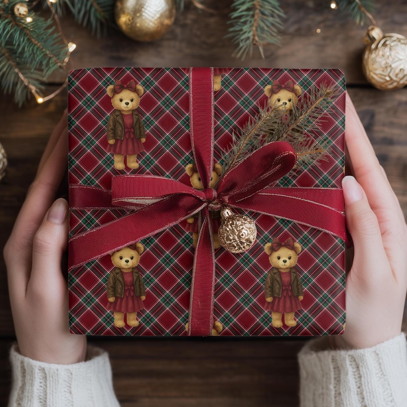 May include: A wrapped gift with red plaid wrapping paper featuring teddy bears in dresses. The gift is tied with a burgundy ribbon and a gold ornament. The gift is held by a person wearing a white sweater.