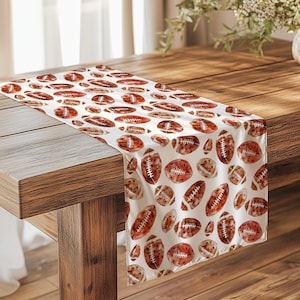 Può includere: Runner da tavolo bianco con un motivo ripetuto di palloni da football in stile acquerello nei toni del marrone e beige. Il runner è drappeggiato su un tavolo di legno.