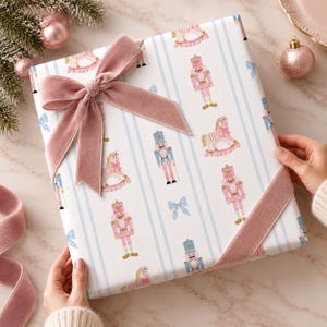 Puede incluir: Una caja de regalo rectangular envuelta en papel blanco con rayas verticales azul claro, con un patrón repetitivo de cascanueces, caballitos de madera y lazos pequeños en rosa y azul, rematada con un gran lazo de terciopelo rosa empolvado.