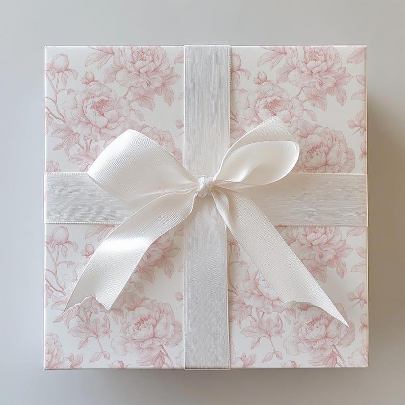 May include: Square gift box with a floral peony pattern in pink and white. The box is tied with a white satin ribbon and bow, creating a classic and elegant presentation. Ideal for special occasions.
