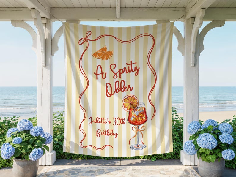 May include: A yellow and white striped banner with the text "A Spritz Older" in red script. The banner also features an illustration of an orange slice and a cocktail. The text "Juliette's 30th Birthday" is also on the banner.
