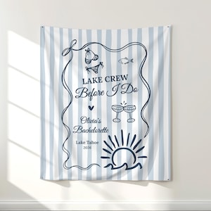 Puede incluir: Un tapiz a rayas azules y blancas con el texto "LAKE CREW Before I Do" e ilustraciones de un bikini, un pez, copas de champán y un sol sobre el agua. También se incluye el texto "Olivia's Bachelorette Lake Tahoe 2026".
