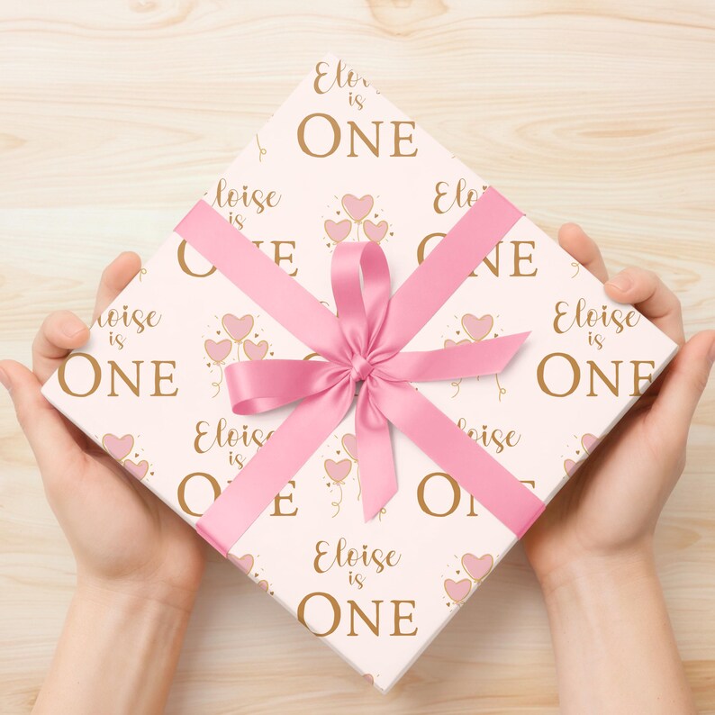 May include: A white gift box with a pink ribbon and a pattern of pink balloons and the text "Eloise is One" in gold lettering.