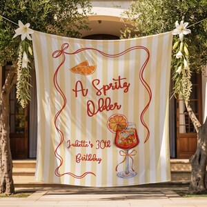 May include: A banner with a yellow and white striped background, featuring the text "A Spritz Older" in red script, along with an illustration of an orange slice and a cocktail. The text "Juliette's 30th Birthday" is also included.