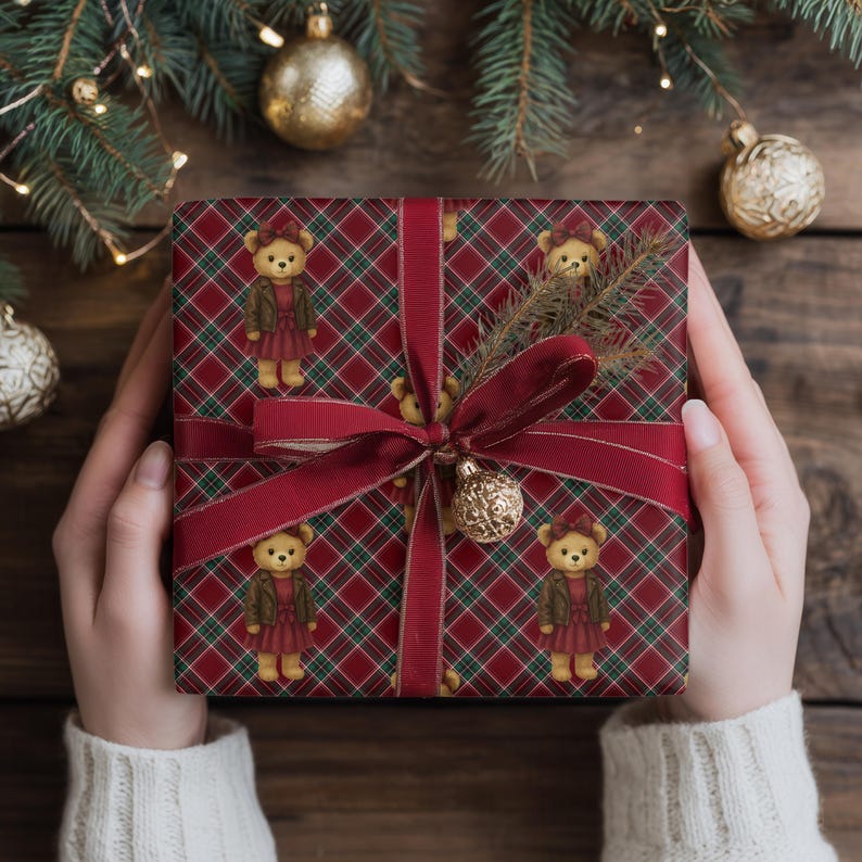 May include: A wrapped gift with a red plaid pattern featuring teddy bears in dresses. The gift is tied with a burgundy ribbon and a gold ornament. The background includes Christmas tree branches and gold ornaments.