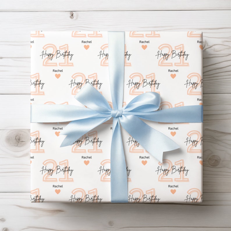 May include: White gift wrap with a light blue ribbon. The wrapping paper has a repeating pattern of the number 21 in peach with the text "Happy Birthday" and the name "Rachel".