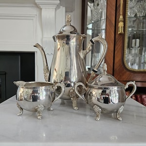 May include: A silver-plated tea service set comprising a tall coffee pot with a lid, a creamer, and a lidded sugar bowl. Each piece features a polished metallic finish, ornate curved handles, and decorative feet with intricate floral detailing.