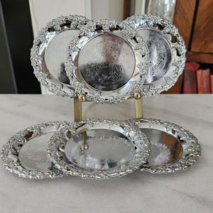 May include: A set of five circular silver-toned metal plates with embossed grape and leaf patterns along the scalloped rims and intricate floral engravings in the centre. The plates are displayed on a gold-toned metal stand with two vertical supports.