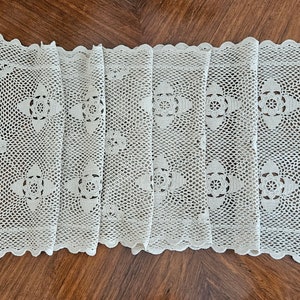 May include: A rectangular off-white crochet table runner featuring a repeating floral motif pattern throughout the centre. The piece is finished with a scalloped edge border and an openwork mesh design, showcasing intricate needlework craftsmanship.