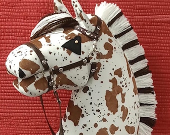May include: A brown and white spotted horse head with a brown leather bridle and reins. The horse head is attached to a wooden stick with a white fringe mane.