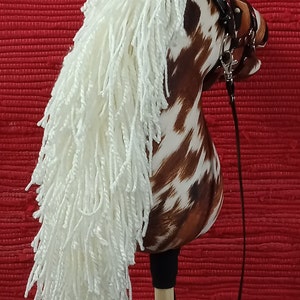 May include: A brown and white spotted horse head with a white mane and a black leather bridle. The horse head is attached to a wooden stick with a black leather rein.