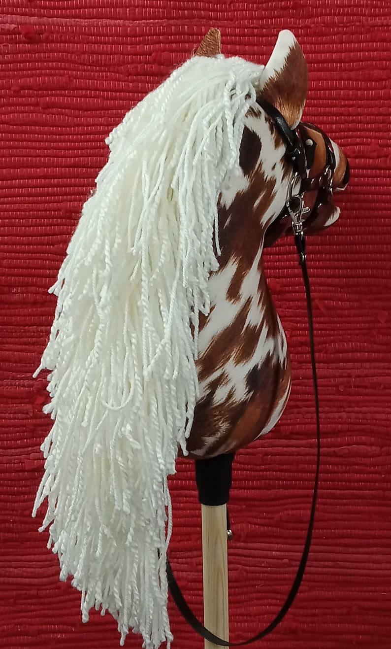 May include: A brown and white spotted horse head with a white mane and a black leather bridle. The horse head is attached to a wooden stick with a black leather rein.