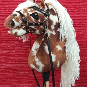 May include: A brown and white spotted horse head with a black leather bridle and white yarn mane and tail. The horse head is attached to a wooden stick with a black leather strap.