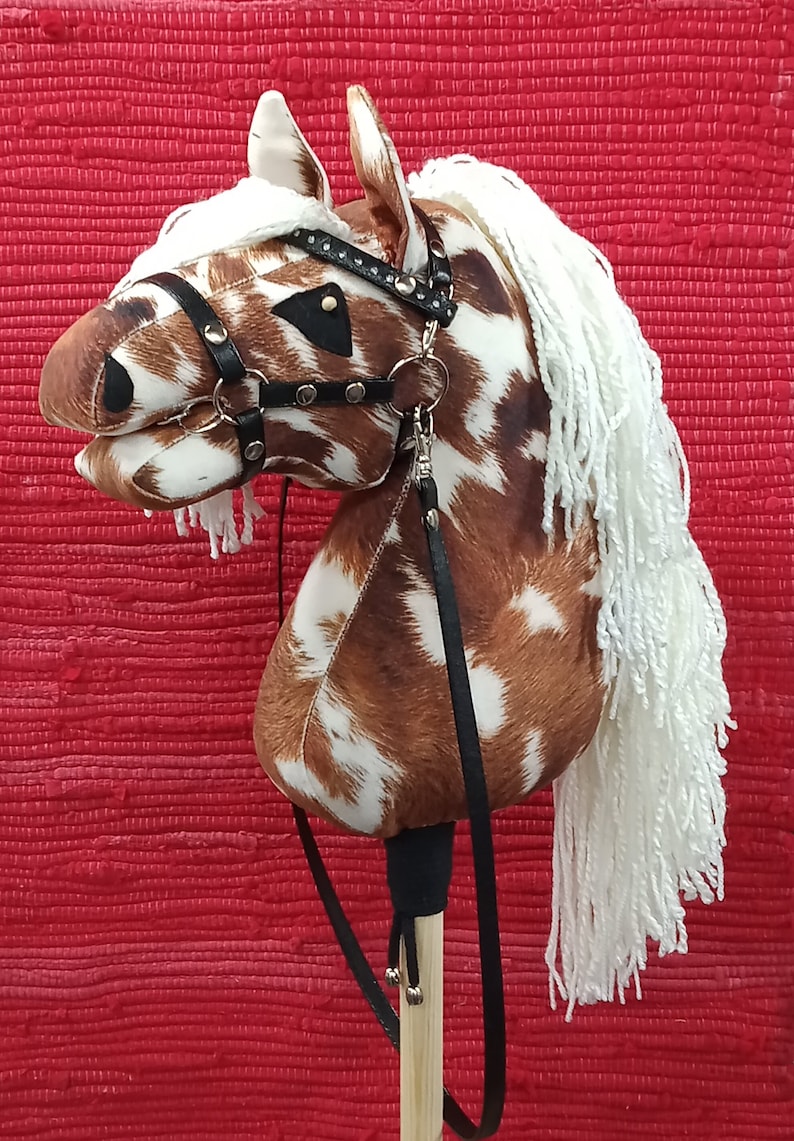 May include: A brown and white spotted horse head with a black leather bridle and white yarn mane and tail. The horse head is attached to a wooden stick with a black leather strap.