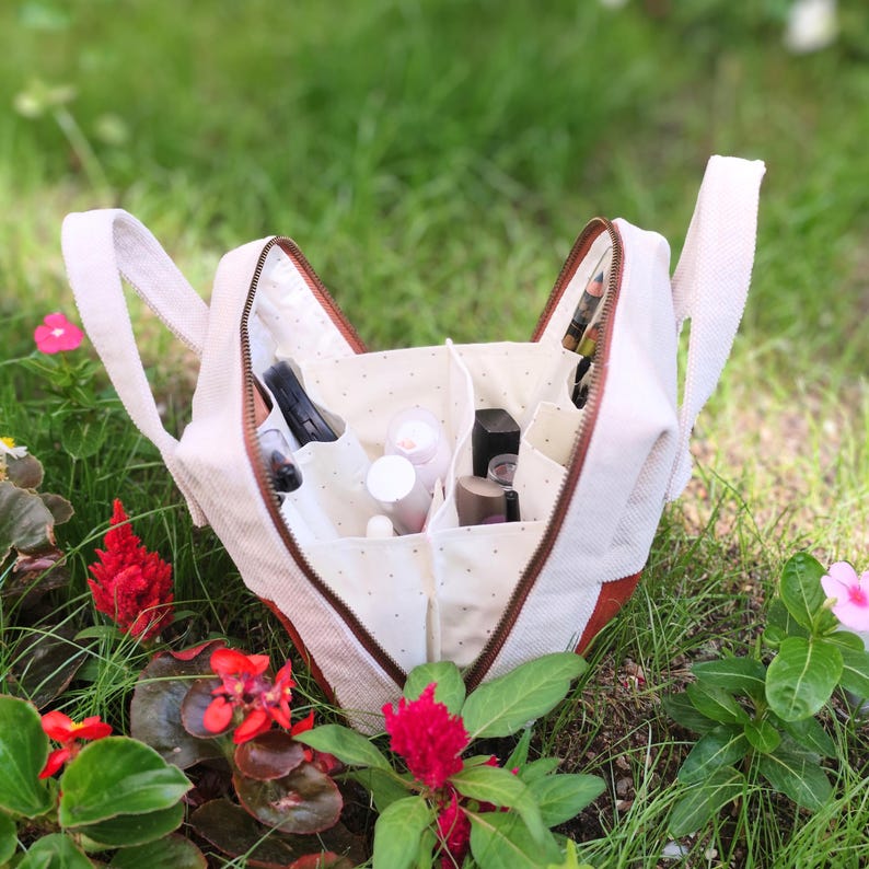 May include: A beige canvas makeup bag with a brown zip and handles, open to show several compartments filled with cosmetics. The bag is set amongst green grass and red and pink flowers.