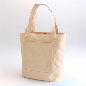 K&ouml;nnte beinhalten: Eine beige Canvas-Tragetasche mit einer gro&szlig;en Tasche auf der Vorderseite. Die Tasche hat zwei lange Henkel.
