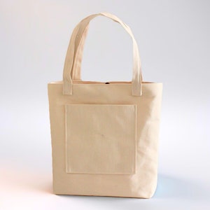 K&ouml;nnte beinhalten: Eine schlichte, cremefarbene Canvas-Tragetasche mit einer gro&szlig;en Tasche auf der Vorderseite. Die Tasche hat zwei lange Henkel.