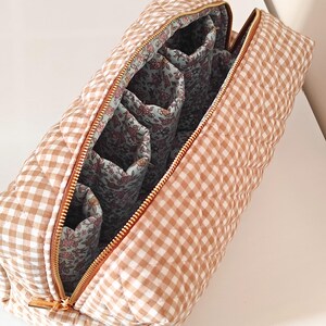 May include: A quilted cosmetic bag with a brown and white gingham pattern. The bag has a gold zipper and a floral patterned interior with multiple compartments. The bag is open, revealing the interior design.