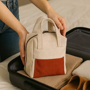 May include: A small, beige travel bag with a rust-coloured front pocket and a brown zip. The bag has two short handles and is placed inside a black suitcase with folded clothing and brown slippers.