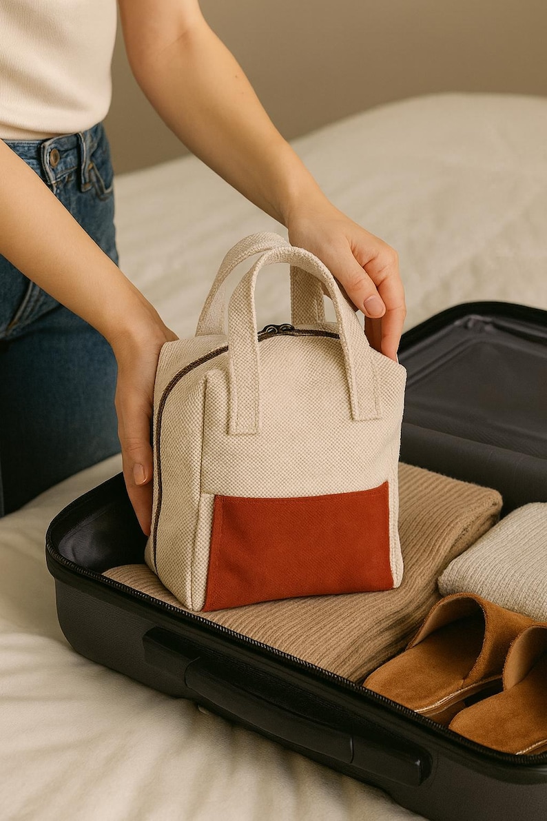 May include: A small, beige travel bag with a rust-coloured front pocket and a brown zip. The bag has two short handles and is placed inside a black suitcase with folded clothing and brown slippers.