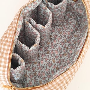 May include: Open view of a beige and white gingham patterned cosmetic bag with a gold zipper. The interior features multiple pockets and a floral print lining in shades of pink, blue, and brown. The bag is designed for organizing beauty products.
