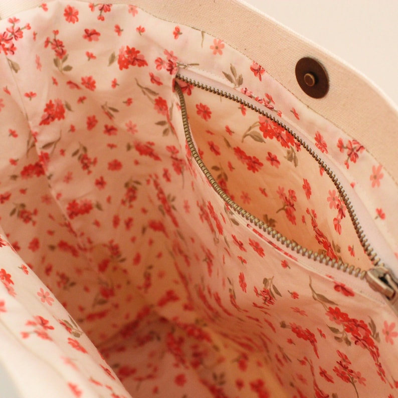 K&ouml;nnte beinhalten: Das Innere einer beigefarbenen Canvas-Tragetasche mit einer Rei&szlig;verschlusstasche. Die Tasche ist mit einem wei&szlig;en Stoff mit einem kleinen roten Blumenmuster gef&uuml;ttert.