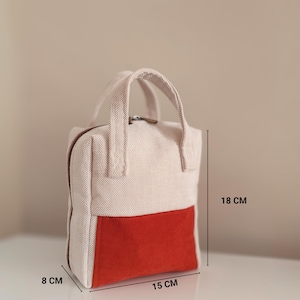 May include: A small, rectangular handbag with a cream-coloured upper section and a vibrant red lower section. The bag, measuring approximately 15 cm wide, 18 cm tall, and 8 cm deep, features a top handle and a zip closure.