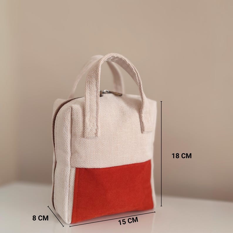May include: A small, rectangular handbag with a cream-coloured upper section and a vibrant red lower section. The bag, measuring approximately 15 cm wide, 18 cm tall, and 8 cm deep, features a top handle and a zip closure.