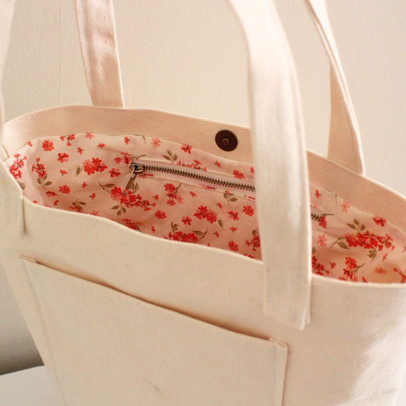 K&ouml;nnte beinhalten: Eine wei&szlig;e Canvas-Tragetasche mit einer Rei&szlig;verschlusstasche und einem floralen Futter. Die Tasche hat zwei Henkel und einen Druckknopfverschluss.