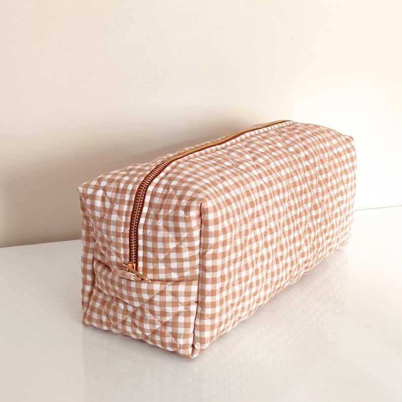 May include: A rectangular, quilted cosmetic bag with a brown and white gingham pattern. The bag features a rose gold zipper closure. The bag is approximately 10 inches long, 5 inches wide, and 5 inches tall.