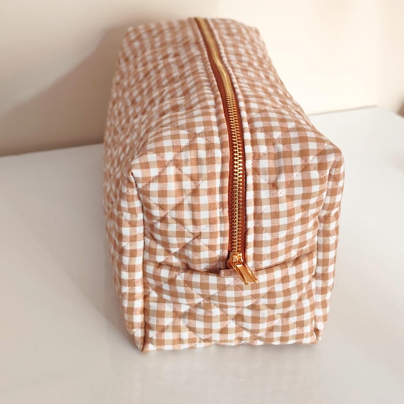 May include: A quilted, rectangular cosmetic bag with a tan and white gingham pattern. The bag features a gold-colored zipper that runs along the top and a matching zipper pull. The bag is designed to hold makeup and other beauty products.