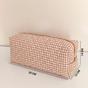 May include: A rectangular, quilted cosmetic bag with a brown and white gingham pattern. The bag has a gold-colored zipper and measures approximately 13 cm high, 14 cm wide, and 35 cm long.