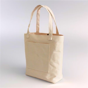 K&ouml;nnte beinhalten: Eine beige Canvas-Tragetasche mit einer gro&szlig;en Tasche auf der Vorderseite. Die Tasche hat zwei Henkel und ist aus robustem Material.