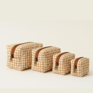 May include: Four beige and white gingham patterned cosmetic bags of varying sizes. Each bag has a brown zipper and a quilted design. The bags are arranged in a row, with the largest on the left and smallest on the right.