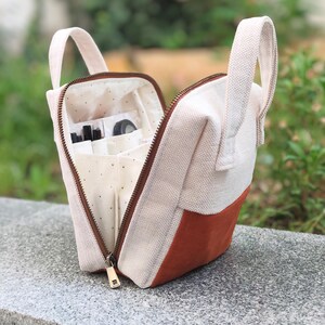 May include: Cream-coloured canvas bag with a brown zip and handle. The bag has a rectangular shape with a section of brown suede-like material on the bottom. The interior has multiple pockets and compartments, with small items visible inside.