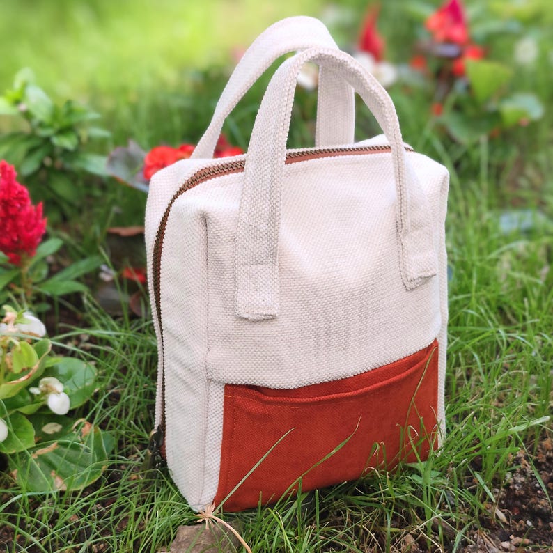 May include: A cream-coloured canvas bag with a rust-coloured front pocket. The bag has two cream-coloured handles and a brown zip. It is set in a grassy area with red flowers in the background. The bag is rectangular.