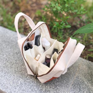 May include: A beige canvas makeup bag with a brown zip and handle. The bag is open, showing multiple compartments filled with various cosmetic products. The interior fabric has a dotted pattern. The bag has a rectangular shape with a brown accent on the bottom.