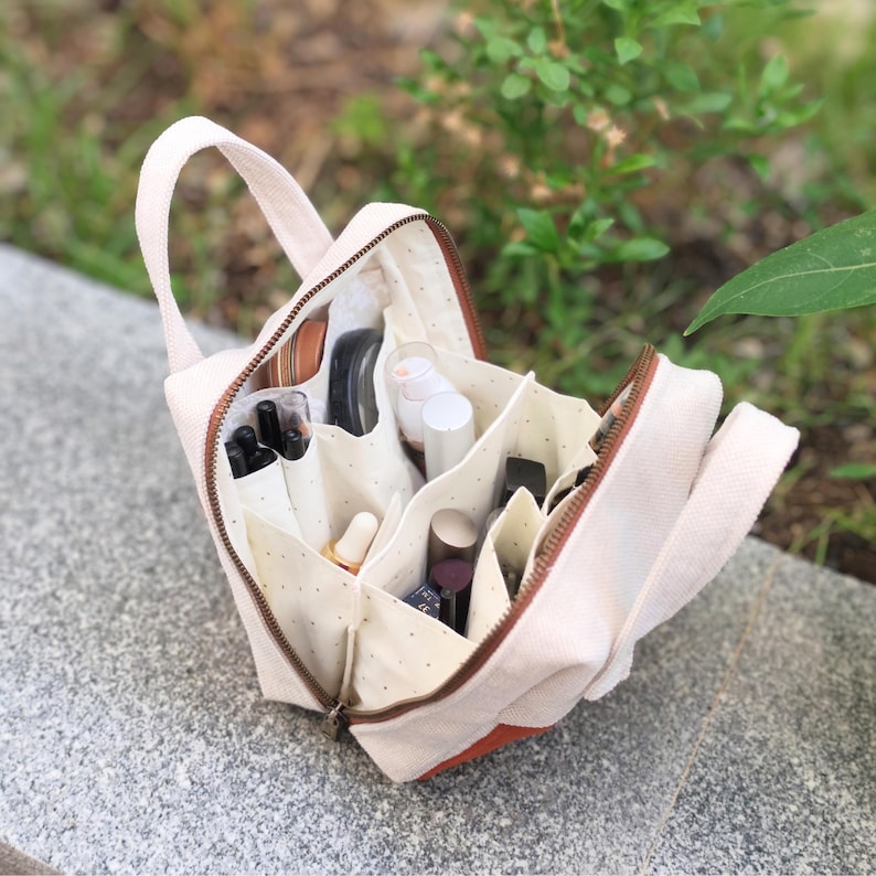 May include: A beige canvas makeup bag with a brown zip and handle. The bag is open, showing multiple compartments filled with various cosmetic products. The interior fabric has a dotted pattern. The bag has a rectangular shape with a brown accent on the bottom.