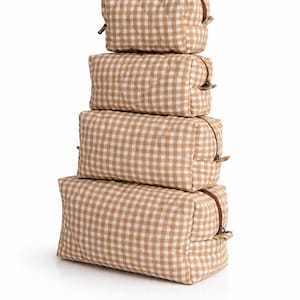 May include: A stack of four rectangular cosmetic bags in varying sizes. Each bag features a tan and white gingham pattern and a brown zipper closure. The bags are arranged vertically against a white background.