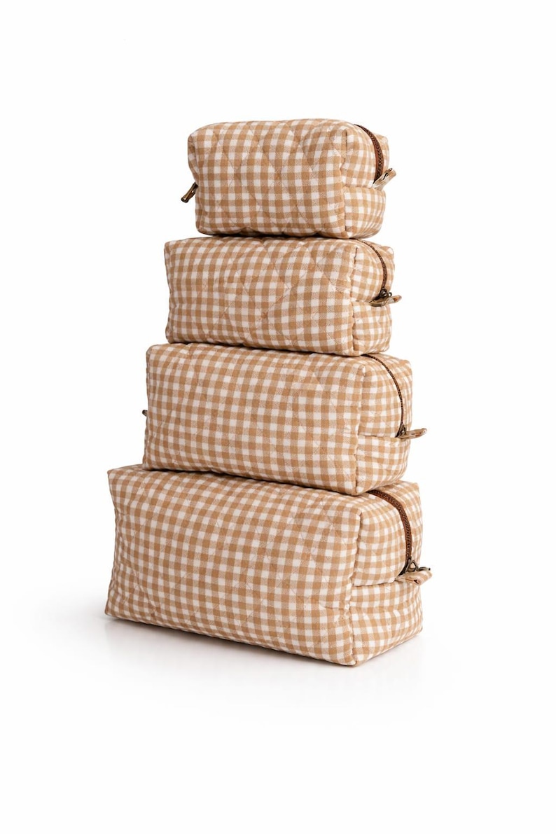 May include: A stack of four rectangular cosmetic bags in varying sizes. Each bag features a tan and white gingham pattern and a brown zipper closure. The bags are arranged vertically against a white background.