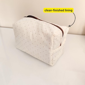 May include: A cream-colored cosmetic bag with a brown zipper and small brown polka dots. The bag has a rectangular shape and a clean-finished lining. The bag is sitting on a white surface.
