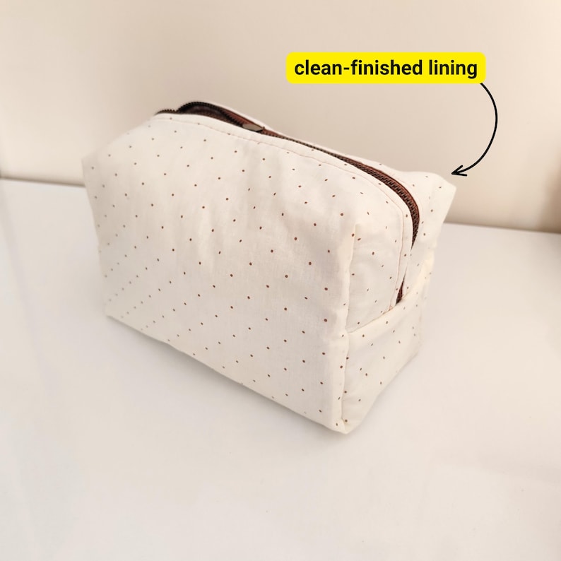 May include: A cream-colored cosmetic bag with a brown zipper and small brown polka dots. The bag has a rectangular shape and a clean-finished lining. The bag is sitting on a white surface.