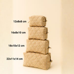 May include: Four stacked, quilted cosmetic bags in varying sizes. The bags feature a tan and white gingham pattern. Measurements are 12x8x8 cm, 16x8x10 cm, 18x10x12 cm, and 22x11x14 cm. Each bag has a zipper closure.