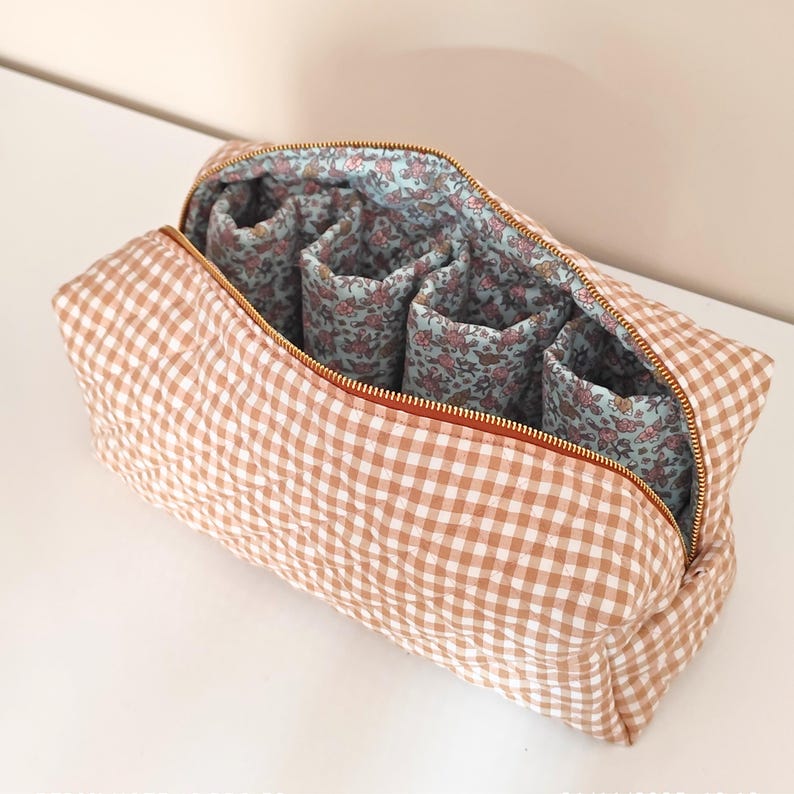 May include: A tan and white gingham cosmetic bag with a gold zipper. The interior features four cylindrical compartments with a floral pattern on a light blue background. The bag is open, revealing the interior design.