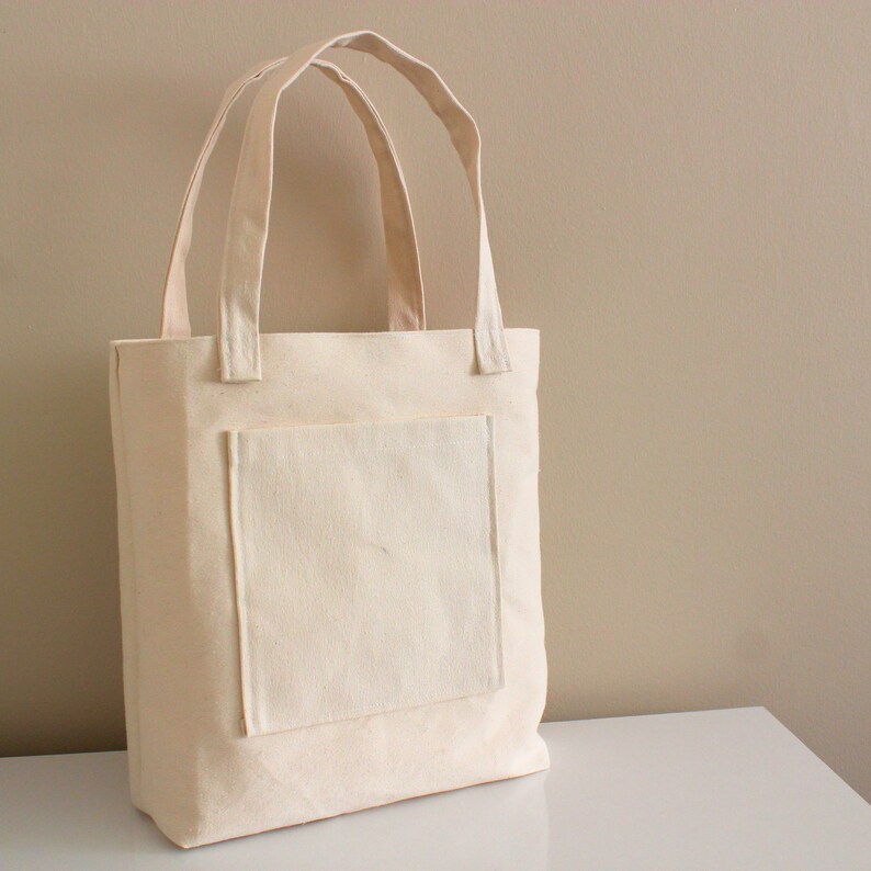 K&ouml;nnte beinhalten: Eine schlichte wei&szlig;e Canvas-Tragetasche mit einer gro&szlig;en Tasche auf der Vorderseite. Die Tasche hat zwei lange Henkel.