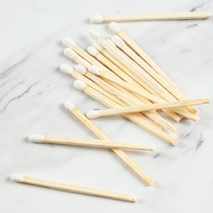May include: A collection of wooden matches with white tips, scattered on a light gray and white marbled surface. The matches are made of natural wood and are arranged in a casual, overlapping pattern.